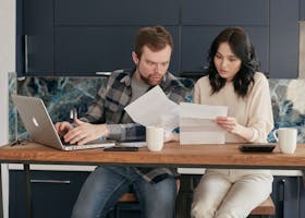 Two coworkers in an office are surprised by the content of a document while working on a laptop.