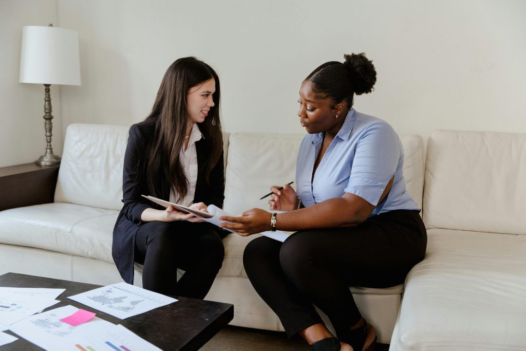 Two businesswomen discussing documents on a sofa in an office setting.