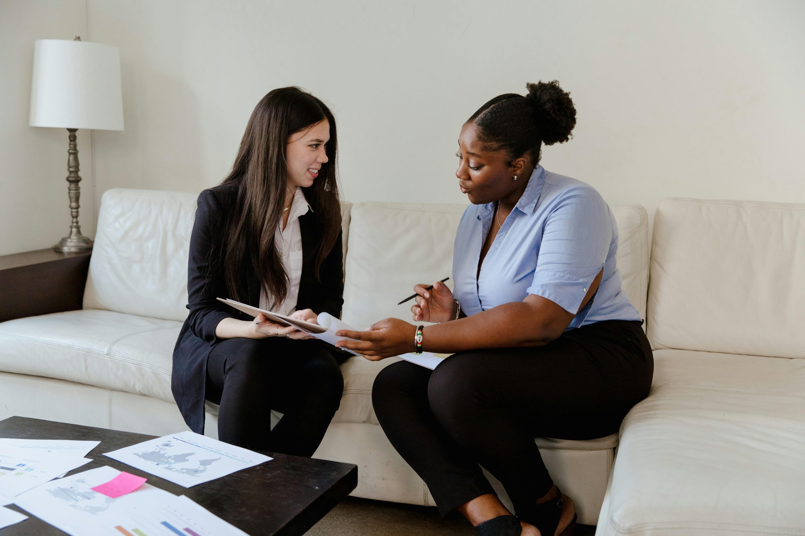 Two businesswomen discussing documents on a sofa in an office setting.